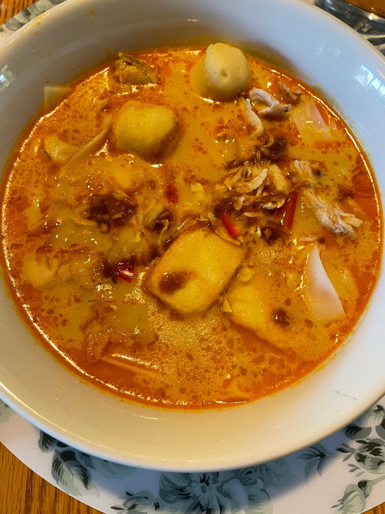 A bowl of bright orange soup with fish balls, shredded chicken, tofu puffs, noodles and chili.