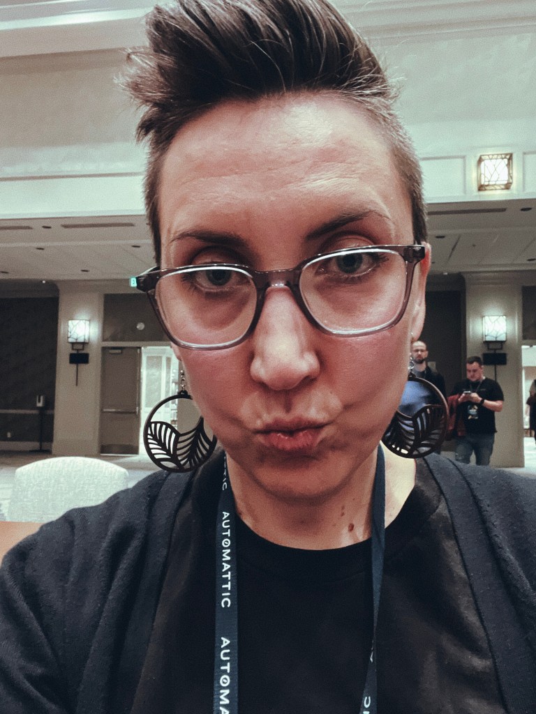 In a conference center making duckface at the camera, wearing big wooden earrings and an Automattic lanyard 