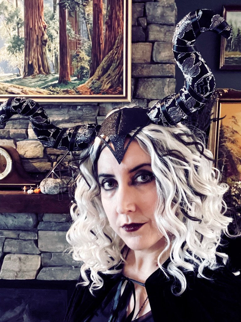 Wearing a curly white wig with black highlights and a black and silver horned headpiece, goth makeup and a black velvet cape 